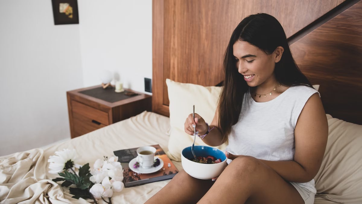 Woman enjoying breakfast in bed with a bowl of granola and fruit, creating a cozy atmosphere.